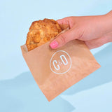 Hand holding a Cereal Milk New York Cookie in Crumbs & Doilies brown paper bag against blue background